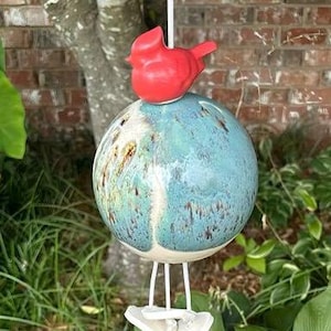 May include: A hanging ceramic bird feeder with a red bird on top of a blue and brown sphere. The feeder is suspended by a white cord and has a white base with three legs.