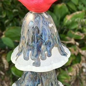 May include: A ceramic wind chime featuring a red bird perched atop a mushroom-shaped bell. The bell has a blue, white, and brown glazed finish. The chime is suspended by a white string, perfect for outdoor decor.