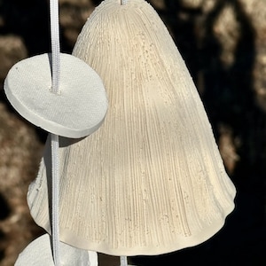 May include: A cream-colored wind chime with a textured, bell-shaped top and several white, circular accents. The chime is suspended by white cords, creating a simple, natural aesthetic. The bell has vertical lines.