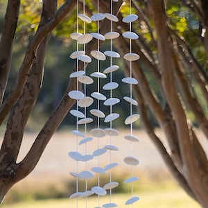 May include: A white ceramic mobile with multiple round discs hanging from a string. The mobile is hanging between two tree branches.