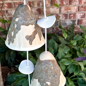 May include: A set of three ceramic wind chimes shaped like mushrooms. The chimes are painted in shades of brown, white, and gray, with a mottled, marbled effect. The chimes are suspended from a white cord.