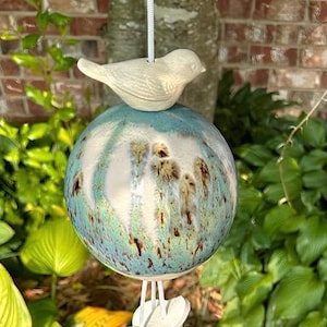 May include: A ceramic birdhouse with a light beige bird perched atop a blue and white speckled sphere. The sphere has brown and green accents. The birdhouse hangs from a white string, with white decorative elements at the bottom.