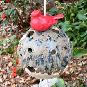 May include: A ceramic birdhouse with a red cardinal perched on top. The birdhouse is spherical, with a speckled gray and blue glaze and several circular openings. It hangs from a white cord, suitable for outdoor decor.