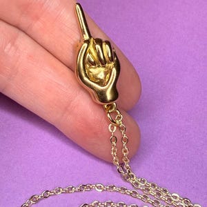 May include: Gold-colored pendant necklace featuring a hand with an extended index finger. The pendant is attached to a delicate gold chain. The background is a solid purple color.