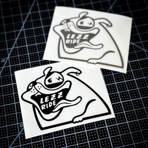 May include: Two black and white stickers featuring a cartoon bulldog with the text "Lezz Ride" on its chest. The stickers are on a black grid background.