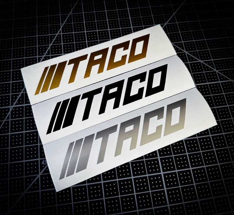 Peut inclure: Trois autocollants en vinyle avec le mot "TACO" dans une police stylis&eacute;e. Les autocollants sont en or, noir et argent.