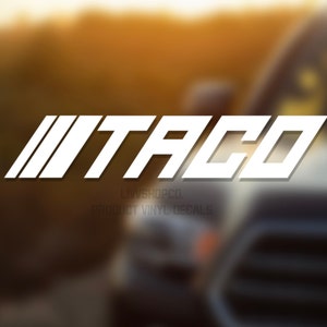 Peut inclure: Autocollant en vinyle blanc avec le mot "TACO" dans une police de caract&egrave;res audacieuse et stylis&eacute;e. L'autocollant est plac&eacute; &agrave; l'arri&egrave;re d'un camion argent&eacute;.