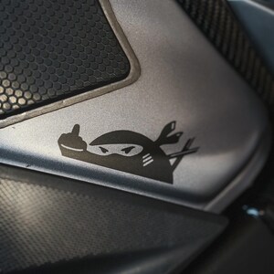 May include: A black and white sticker of a ninja giving the middle finger, on a silver motorcycle part with a black honeycomb pattern.