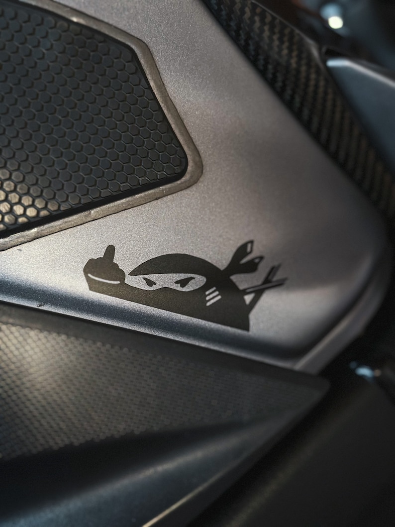 May include: A black and white sticker of a ninja giving the middle finger, on a silver motorcycle part with a black honeycomb pattern.