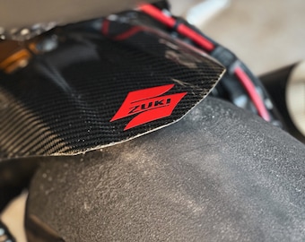 May include: Close-up of a black carbon fiber motorcycle fender with a red "ZUKI" logo.