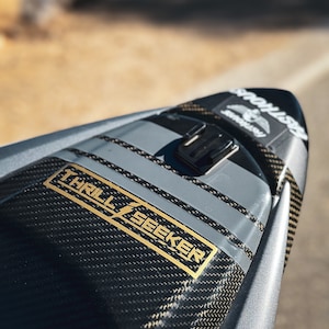 May include: A close-up of a black and gray motorcycle seat with a carbon fiber pattern. The seat has a gray stripe down the center and a gold sticker that says "THRILL SEEKER".
