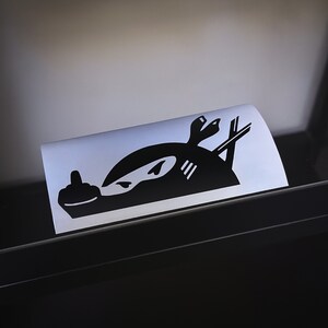May include: Black and white sticker of a ninja giving the middle finger. The ninja is wearing a mask and has a katana sword behind them.