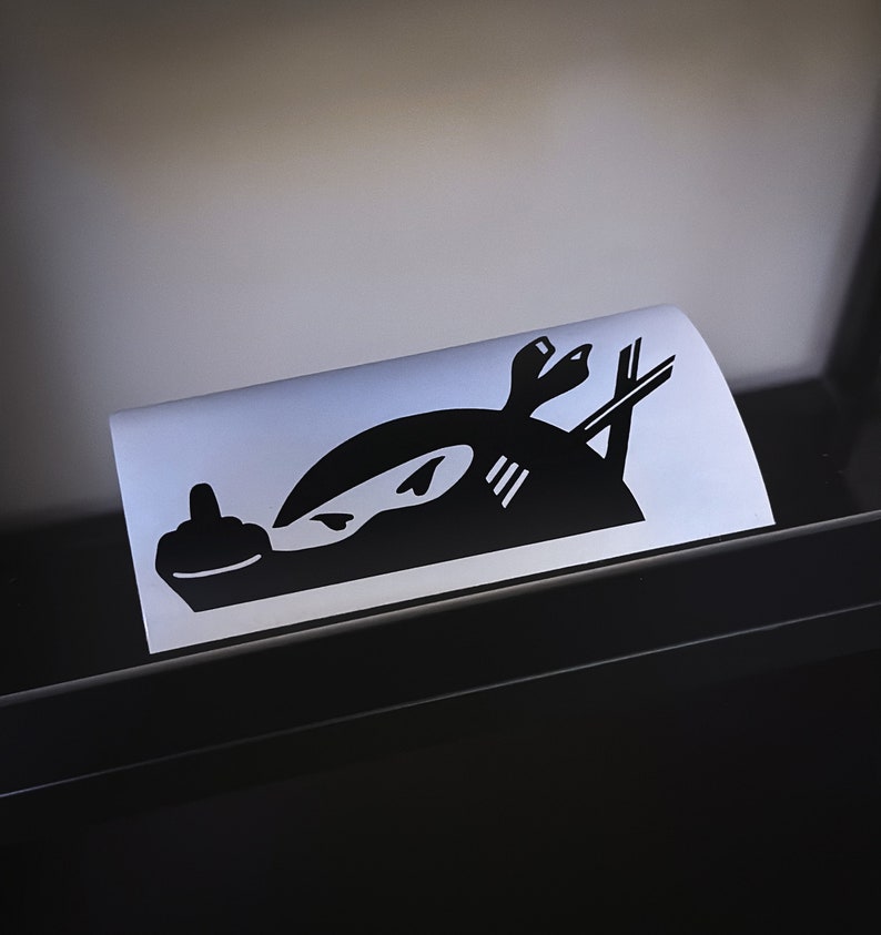 May include: Black and white sticker of a ninja giving the middle finger. The ninja is wearing a mask and has a katana sword behind them.