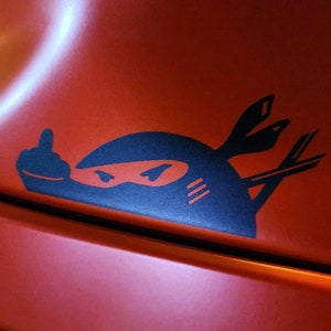 May include: A black and white sticker of a ninja giving the middle finger. The ninja is wearing a black mask and has a white stripe on its forehead. The sticker is on a red surface.