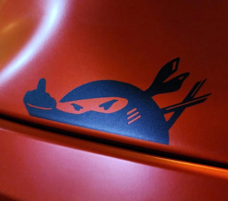 May include: A black and white sticker of a ninja giving the middle finger. The ninja is wearing a black mask and has a white stripe on its forehead. The sticker is on a red surface.