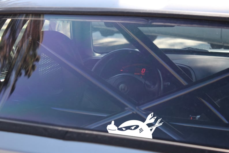 May include: A white sticker of a cartoon character with a middle finger on a car window. The character is wearing a helmet and has a sword in its hand.