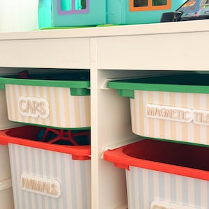 May include: A white storage unit with four plastic bins featuring striped patterns and white labels. The bins are labeled Cars, Magnetic Tiles, Animals, and Crafts. The top left bin is green, the top right is green, and the bottom two are red.