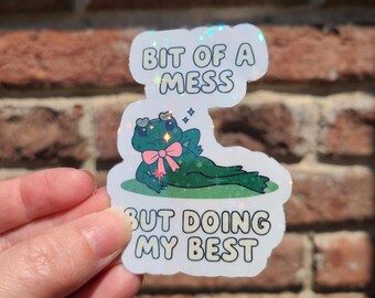 May include: A holographic sticker featuring a cartoon frog with a pink bow, lounging on grass. The sticker reads "Bit of a Mess But Doing My Best" in white text with a blue outline. The background is a brick wall.