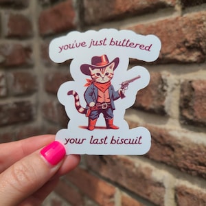 May include: A sticker featuring a cartoon cat dressed as a cowboy, holding a gun. The cat wears a hat, red neckerchief, and boots. Text reads "you've just buttered" and "your last biscuit."