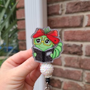 May include: A green caterpillar badge reel with a red bow, glasses, and a book. The badge reel has a glittery border and a white, sparkly bead. The badge reel is held by a clear plastic clip.