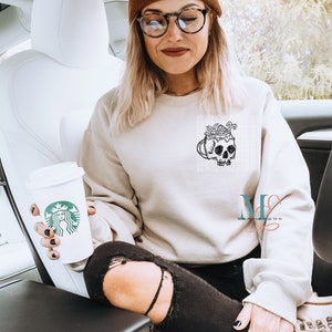 May include: A woman wearing a beige sweatshirt with a black skull graphic and a brown beanie sits in a car holding a white Starbucks cup.