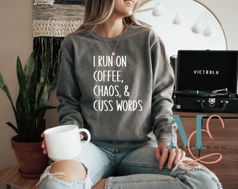 May include: A gray sweatshirt with the text "I run on coffee, chaos, & cuss words" printed on the front. The sweatshirt is being worn by a person holding a white coffee mug.