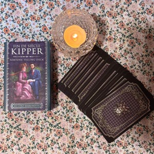 Puede incluir: Una baraja de adivinación titulada "Fin de Siècle Kipper" y una vela encendida en un portavelas de cristal descansan sobre una superficie con estampado floral. La caja de la baraja muestra una pareja con ropa de época. Una baraja de cartas está abanicada junto a la caja.