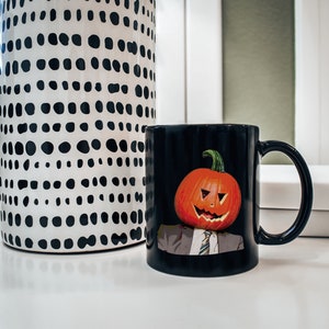 May include: Black coffee mug with a pumpkin face design. The pumpkin has a carved smile and a tie. The mug is on a white surface next to a black and white polka dot vase.