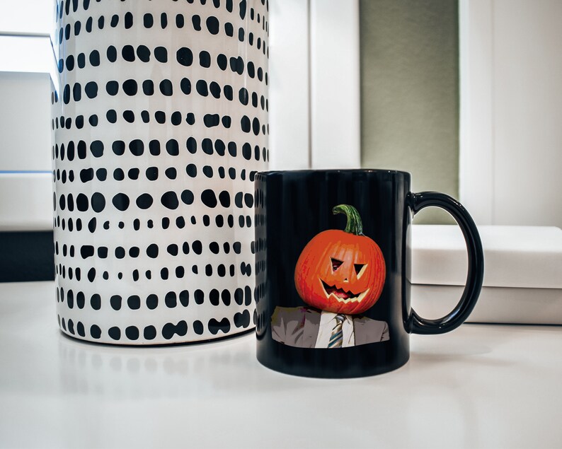 May include: Black coffee mug with a pumpkin face design. The pumpkin has a carved smile and a tie. The mug is on a white surface next to a black and white polka dot vase.