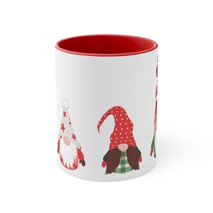 May include: White ceramic mug with a red rim. The mug features a design of three gnomes with red and white hats and green and white clothing.
