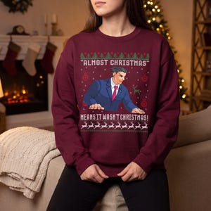 May include: A maroon long-sleeve sweatshirt featuring a graphic of a man in a blue suit and red tie. The text reads "ALMOST CHRISTMAS" and "MEANS IT WASN'T CHRISTMAS!" surrounded by a pattern of green trees, red ornaments, and white reindeer.