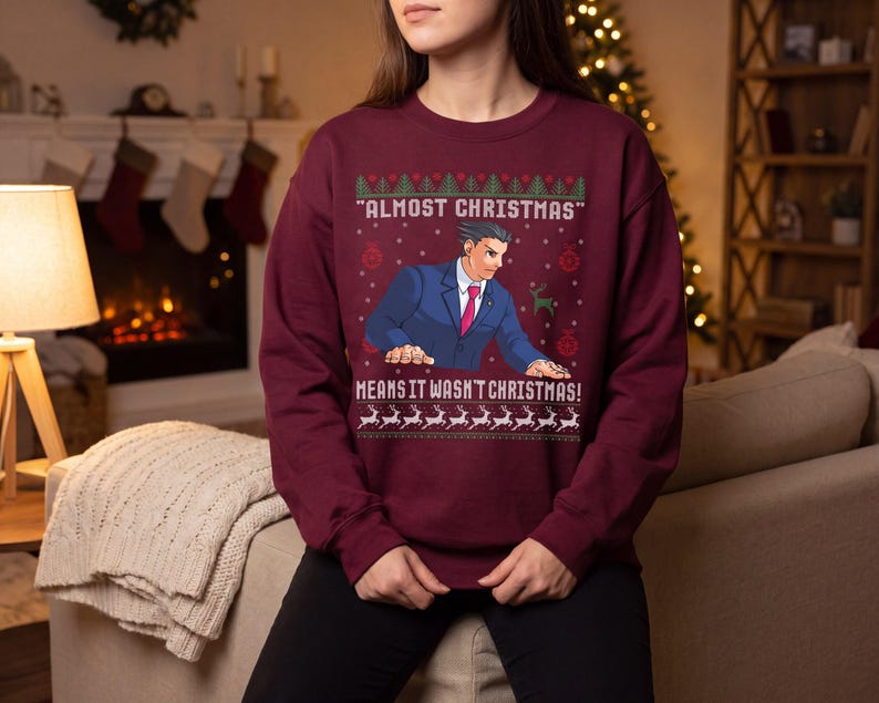 May include: A maroon long-sleeve sweatshirt featuring a graphic of a man in a blue suit and red tie. The text reads "ALMOST CHRISTMAS" and "MEANS IT WASN'T CHRISTMAS!" surrounded by a pattern of green trees, red ornaments, and white reindeer.