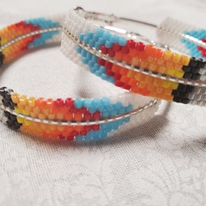 May include: Two beaded hoop earrings with a colourful geometric pattern in shades of white, black, blue, yellow, orange and red.