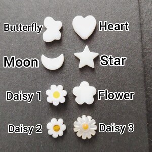 May include: A collection of white, pearl-like embellishments in various shapes: butterfly, heart, moon, star, and three daisy and flower designs. Each shape is labeled with its name in white text. The embellishments are arranged on a dark gray surface.