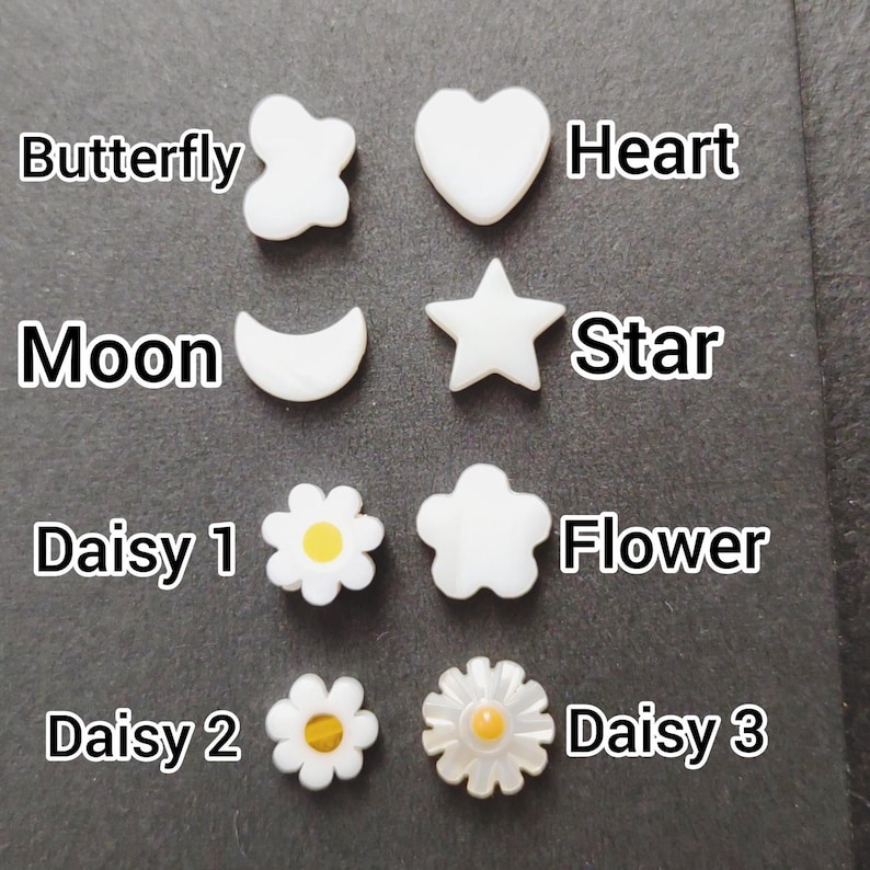 May include: A collection of white, pearl-like embellishments in various shapes: butterfly, heart, moon, star, and three daisy and flower designs. Each shape is labeled with its name in white text. The embellishments are arranged on a dark gray surface.