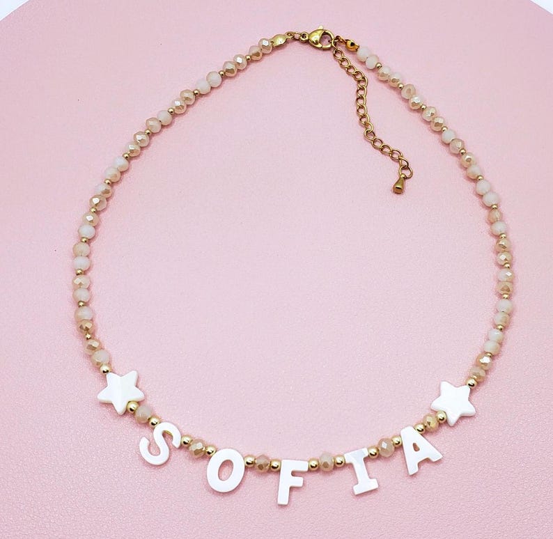 May include: A beaded necklace with the name "SOFIA" spelled out in white letters. The necklace features small, light-colored beads and gold-colored accents. Two white star-shaped charms flank the name. The necklace is displayed on a pink surface.
