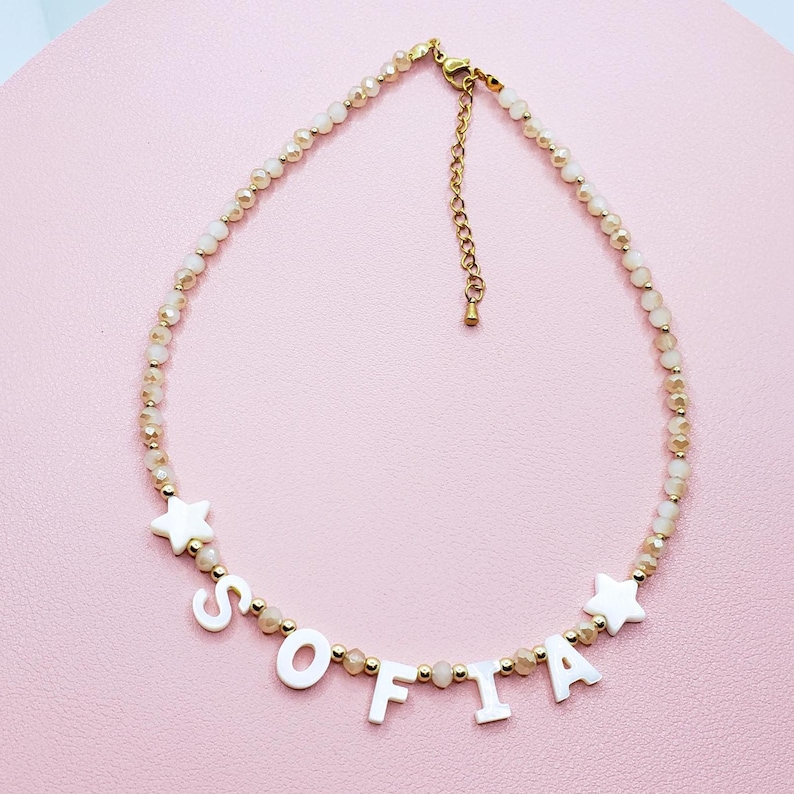 May include: A beaded necklace with the name "SOFIA" spelled out in white letters. The necklace features small, light-colored beads and gold-colored accents. Two white star-shaped charms flank the name. The necklace is displayed on a pink surface.