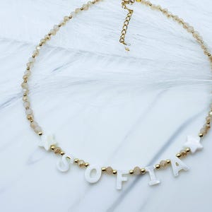 May include: A delicate necklace with the name "SOFIA" spelled out in white letters. The necklace features small, light brown beads and gold accents. Two white star-shaped charms are also included in the design. The necklace is on a white surface.