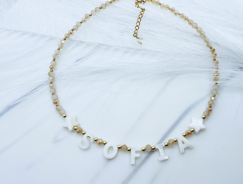 May include: A delicate necklace with the name "SOFIA" spelled out in white letters. The necklace features small, light brown beads and gold accents. Two white star-shaped charms are also included in the design. The necklace is on a white surface.