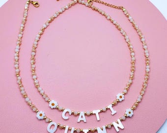 May include: Two beaded necklaces featuring alternating small translucent beige and white beads with gold-tone spacers. One necklace spells CATI and the other spells QUINN using white letter beads, each accented with small white and orange flower charms.