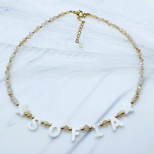 May include: A gold-toned necklace with the name "SOFIA" spelled out in white letter beads. The necklace features small, light brown beads and gold accents. The clasp and chain are gold.