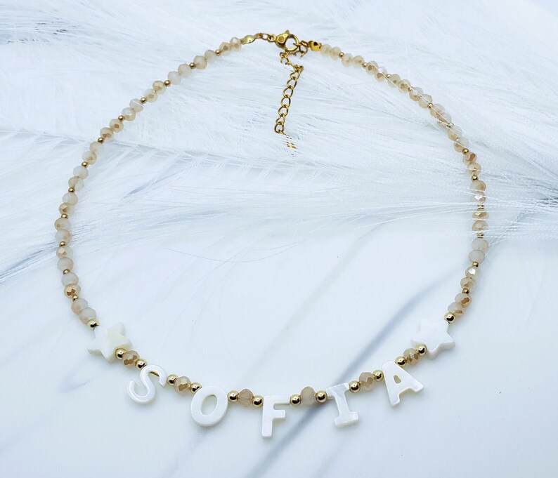 May include: A gold-toned necklace with the name "SOFIA" spelled out in white letter beads. The necklace features small, light brown beads and gold accents. The clasp and chain are gold.