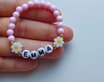 May include: A pink beaded bracelet with the name "EMMA" spelled out in white letter beads. The bracelet features daisy-shaped accents with yellow centers and gold-colored beads. A charming accessory.