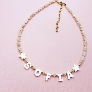 May include: A delicate necklace with the name "SOFIA" spelled out in white letters. The necklace features small, light-colored beads and gold accents, including star-shaped charms. The background is a soft pink.