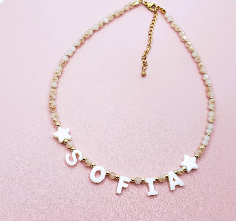 May include: A delicate necklace with the name "SOFIA" spelled out in white letters. The necklace features small, light-colored beads and gold accents, including star-shaped charms. The background is a soft pink.