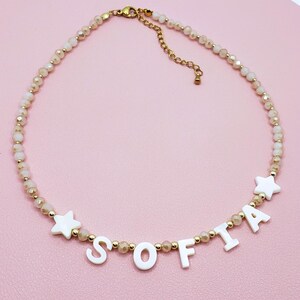 May include: A beaded necklace with the name "SOFIA" spelled out in white letters. The necklace features small, light-colored beads and gold-colored accents. Two white star-shaped charms are also included in the design. The necklace is displayed on a pink surface.
