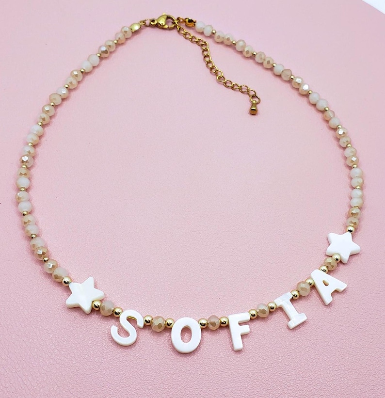 May include: A beaded necklace with the name "SOFIA" spelled out in white letters. The necklace features small, light-colored beads and gold-colored accents. Two white star-shaped charms are also included in the design. The necklace is displayed on a pink surface.