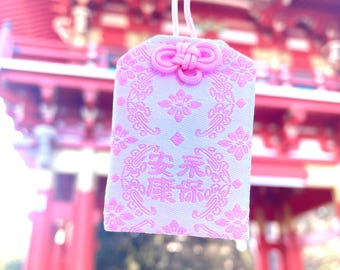 Puede incluir: Un amuleto omamori japonés rosa con un patrón floral y el texto "安康保" en japonés. El amuleto cuelga de un cordón blanco, con un nudo decorativo en la parte superior. El fondo es una vista borrosa de un templo japonés rojo.