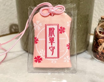 Puede incluir: Un amuleto omamori japonés rosa con un diseño rectangular. El amuleto presenta un texto enmarcado en rojo y detalles florales. Una cuerda y un lazo rosas están unidos en la parte superior. El amuleto está encerrado en una funda de plástico transparente.