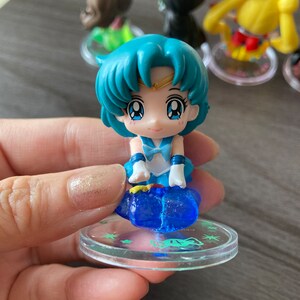 Puede incluir: Una peque&ntilde;a figura de un personaje con cabello azul y un atuendo azul, de pie sobre una base transparente. La figura tiene ojos grandes y forma parte de una colecci&oacute;n de figuras similares. Otras figuras son visibles en el fondo.
